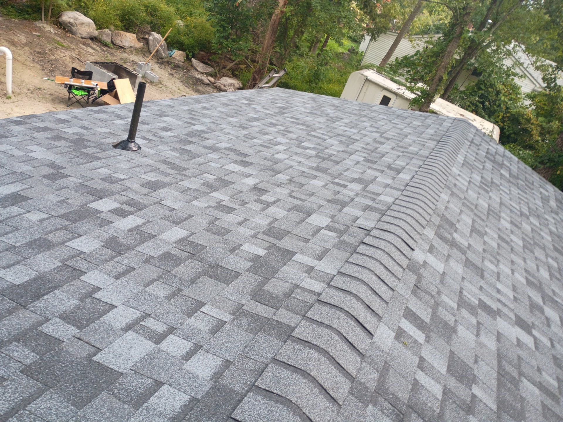 Gray asphalt shingle roof, with a vent pipe and curved ridge, in an outdoor setting.