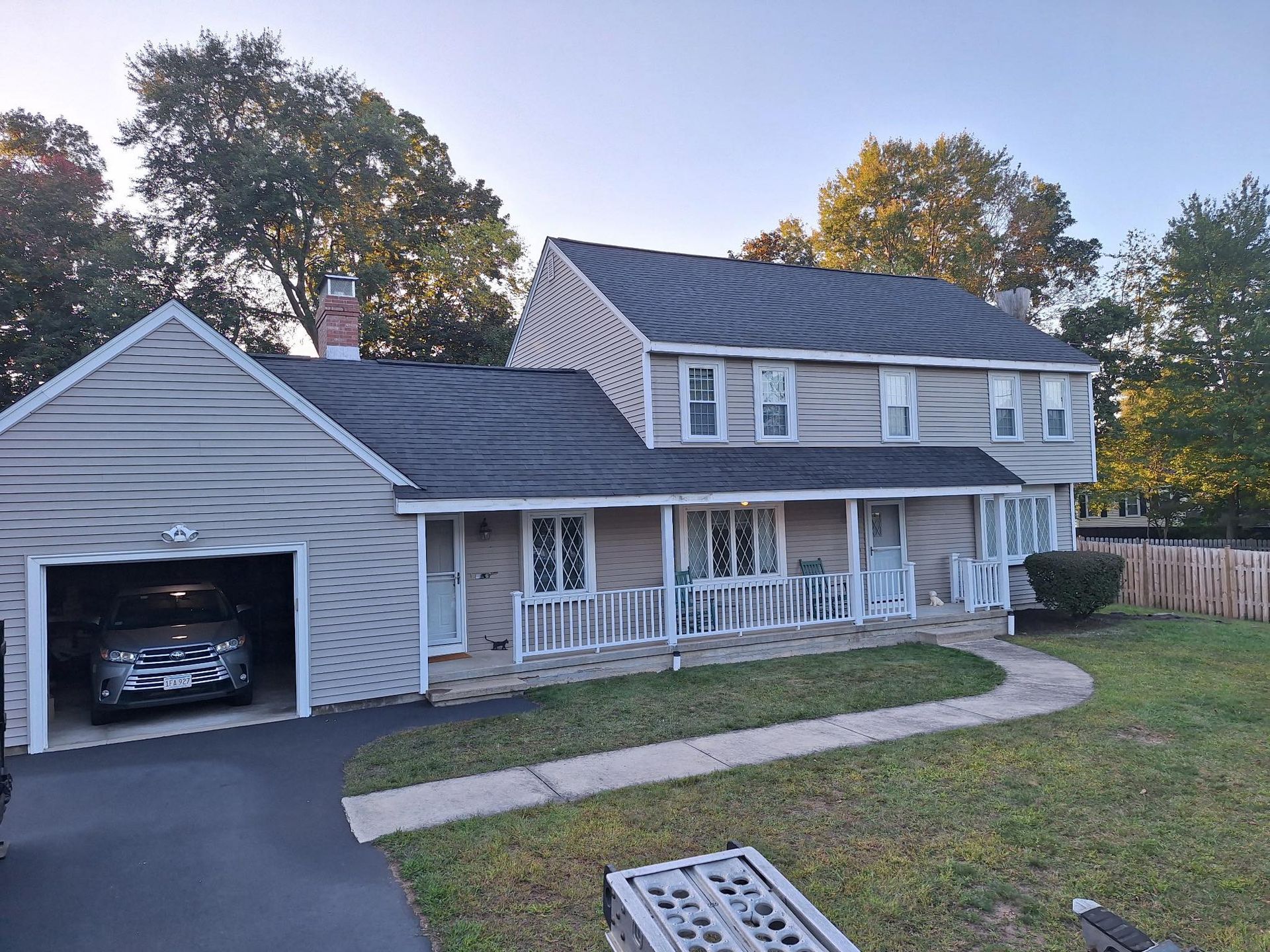 Two-story beige house with attached garage, dark roof, white porch, and a car parked inside.