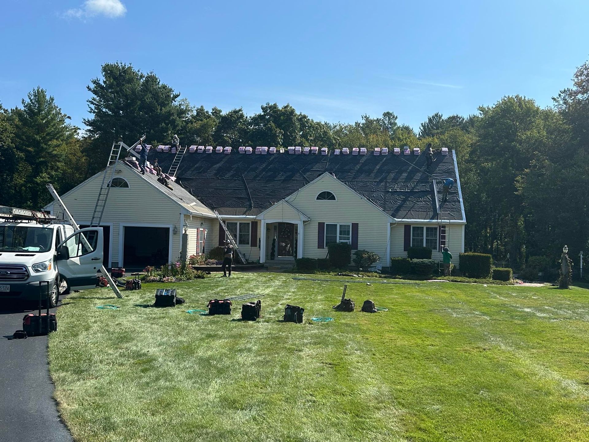 Workers replacing shingles on a light yellow house roof. Ladders and tools are present. Green lawn with trees.