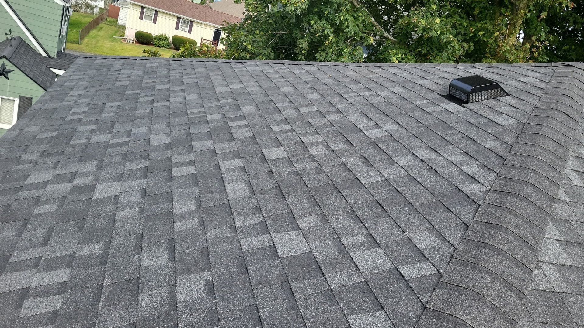 Dark gray asphalt shingle roof with a vent. Houses and trees in the background.