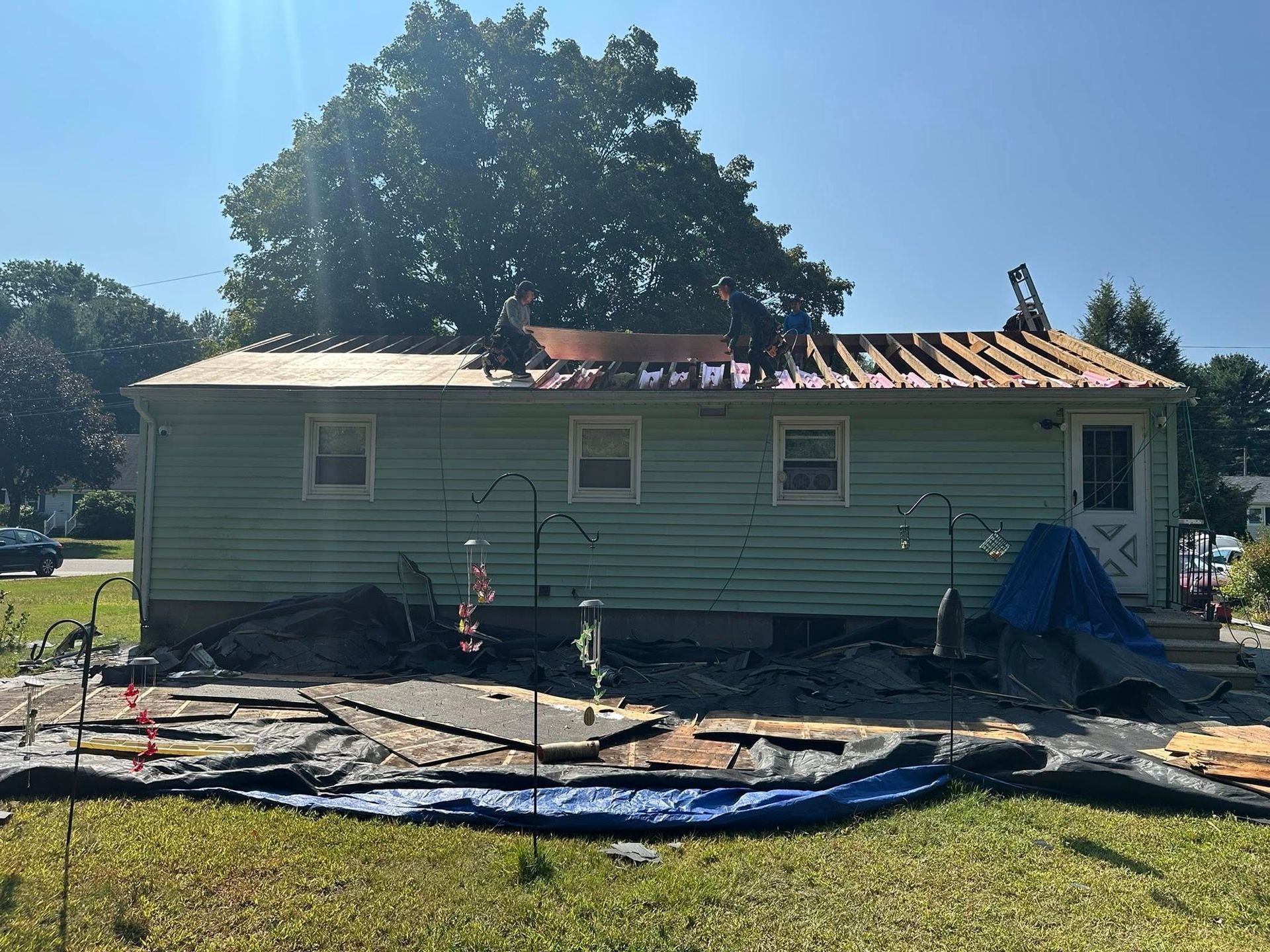 Roofers working on a house with a partially removed roof; green siding, blue tarp on the ground, sunny day.