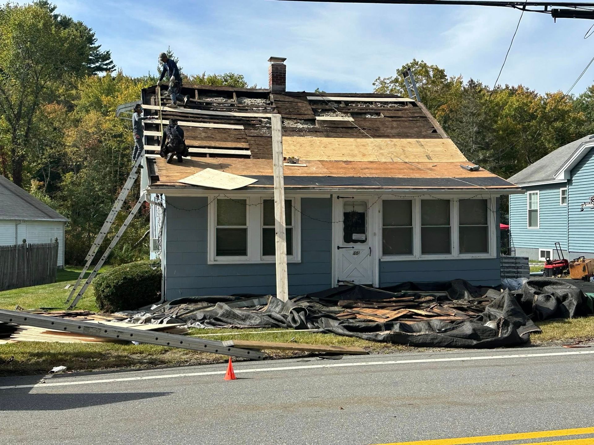 House under construction; roof partially removed, workers on the roof. Blue house, setting is outdoors on a sunny day.