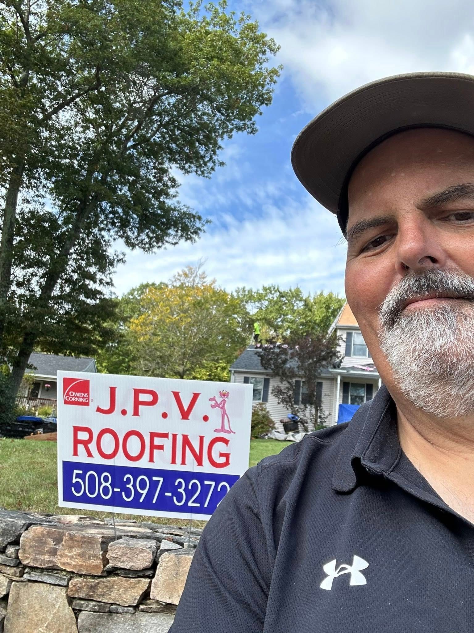 Man smiles near a J.P.V. Roofing sign in front of a house and trees. Blue sky.