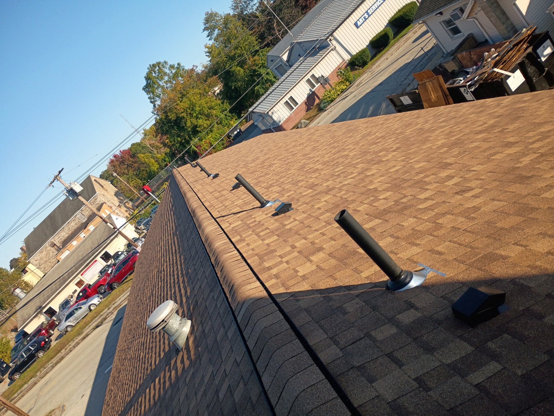 Brown shingled roof with vents and pipes against a clear, sunny sky. Buildings and street visible in background.