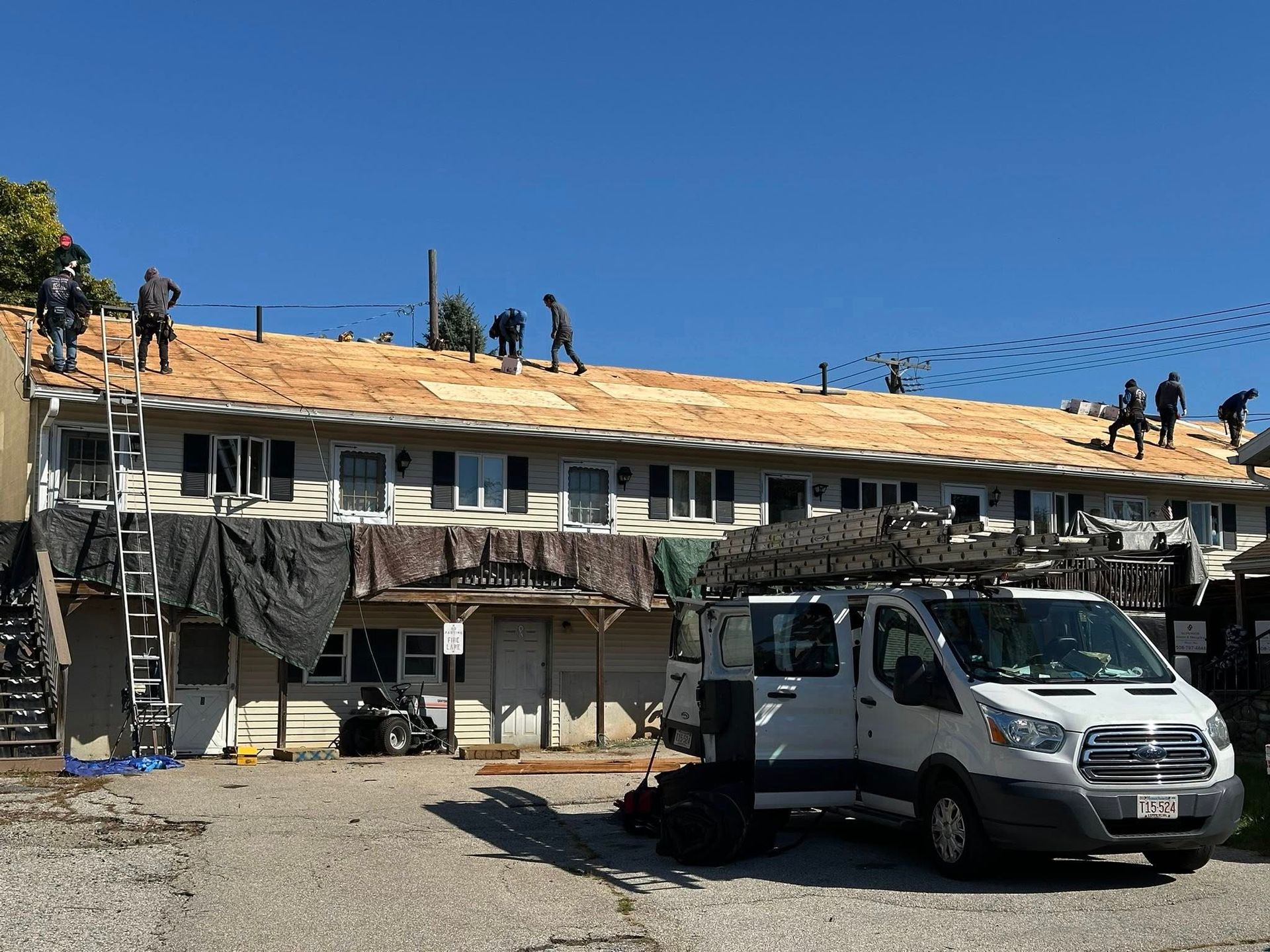 Roofers working on an apartment building, sky overhead. White van parked in front.