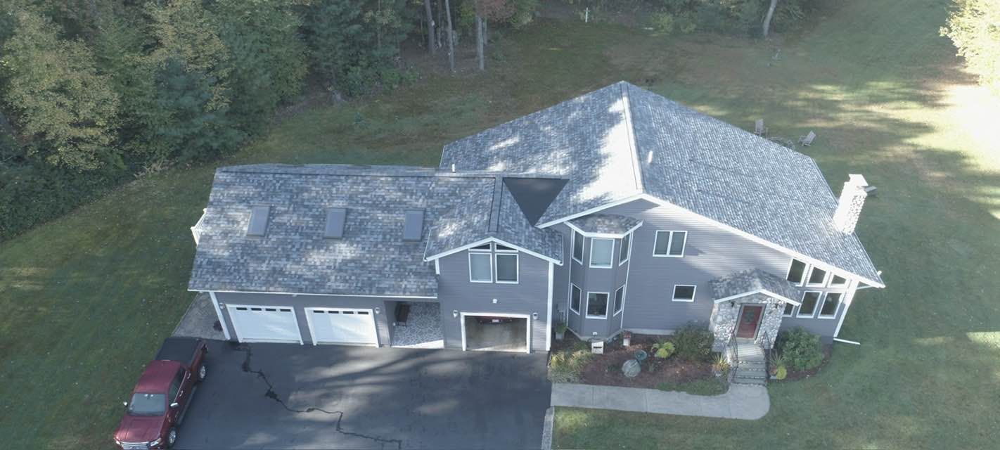 Aerial view of a gray house with a black driveway, surrounded by trees and green grass.