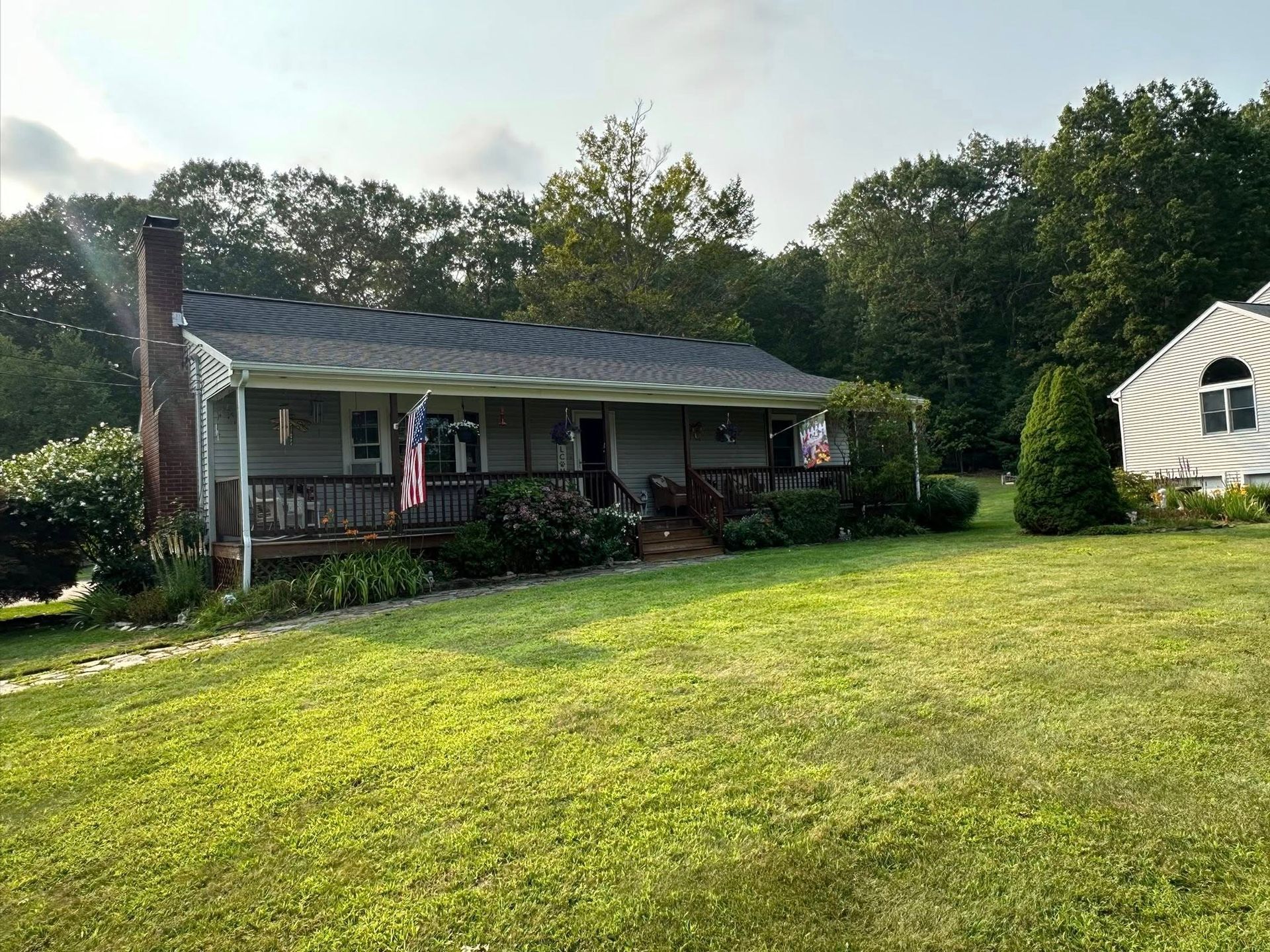 A gray ranch home with a porch, chimney, and American flags sits on a grassy hill with trees in the background.