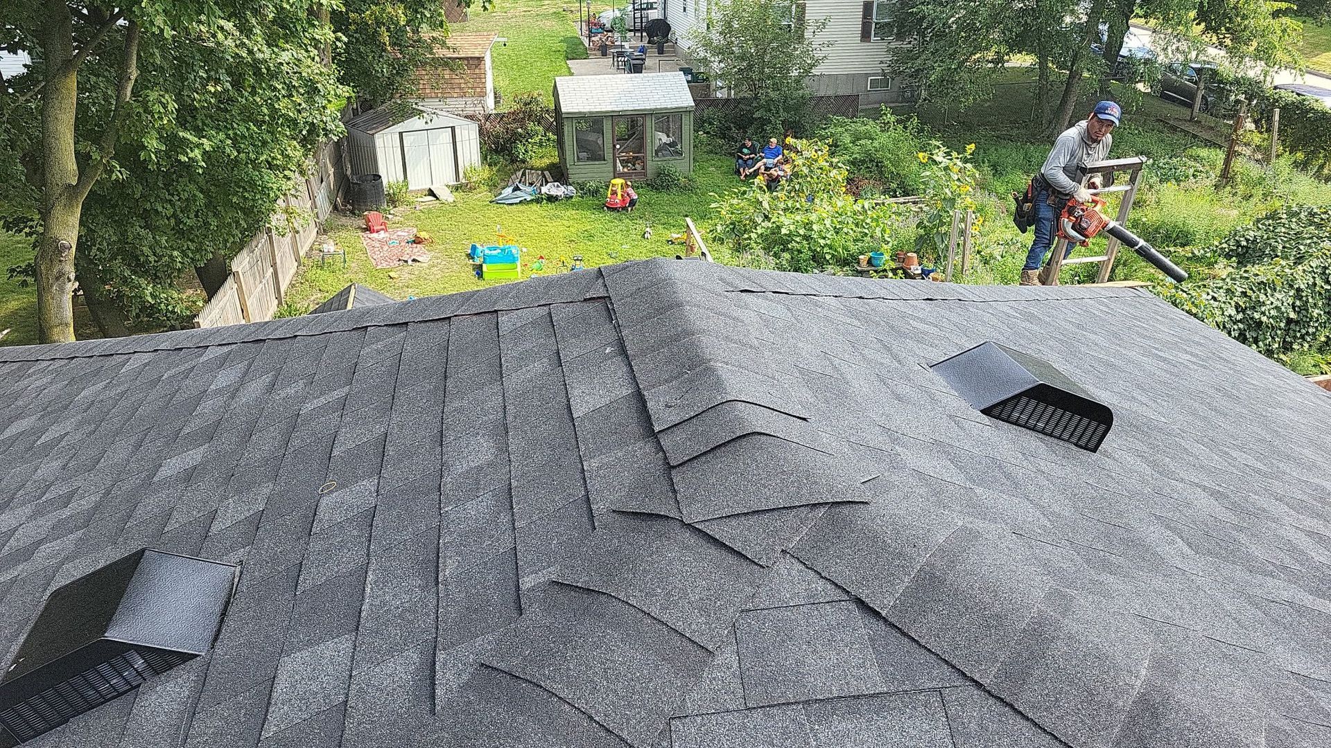 Roofer on a gray shingle roof, using a blower. Green yard, trees, and buildings are in the background.