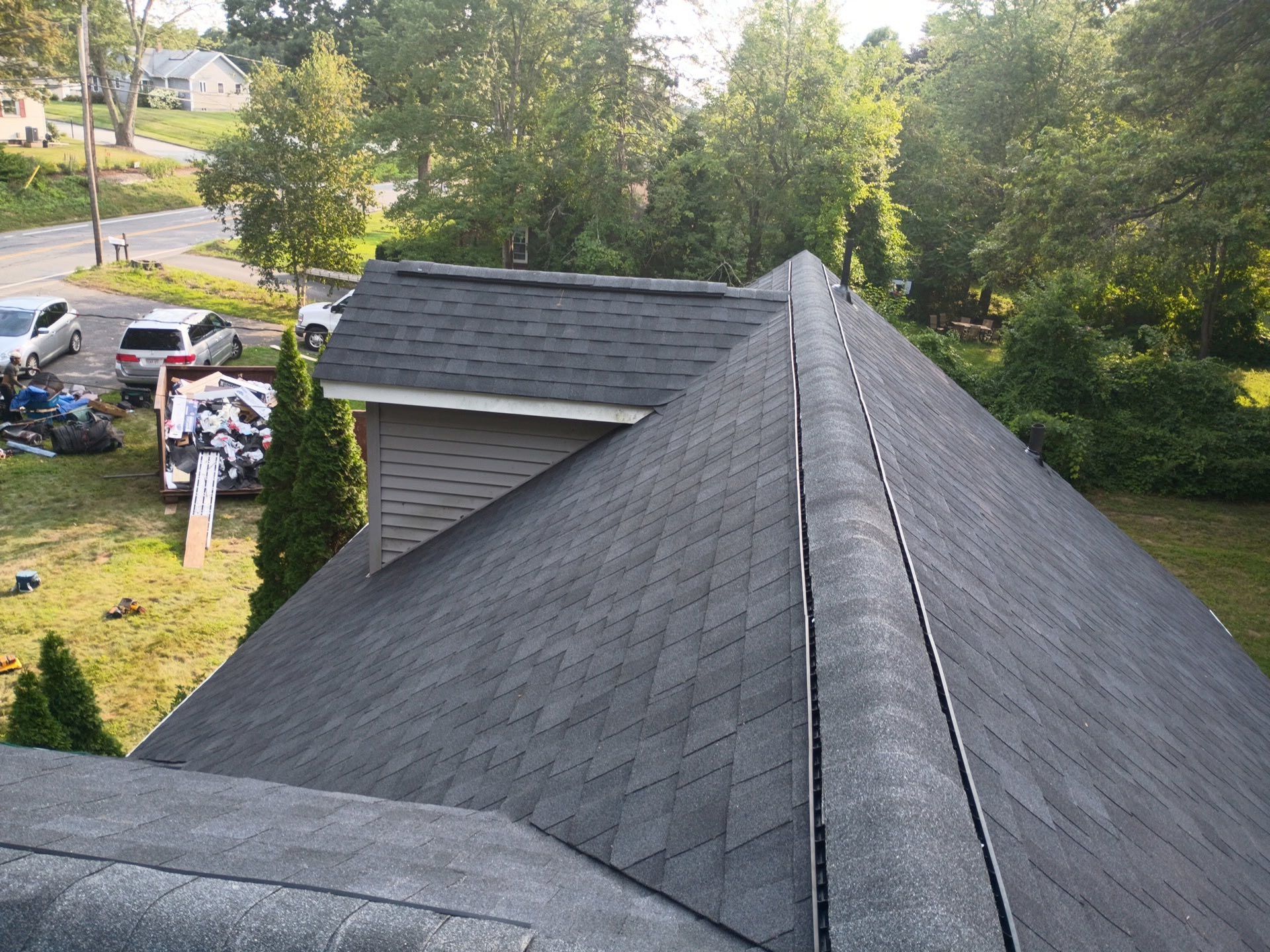 View of a dark gray shingled roof from above, looking towards a street with parked cars and trees.