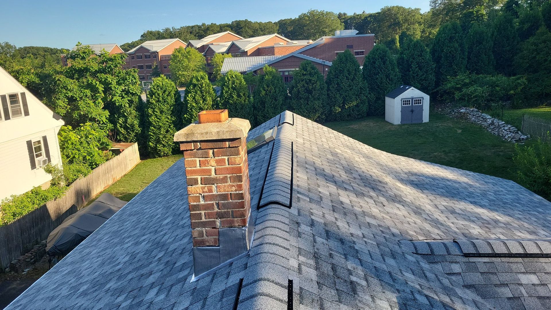 Rooftop view with brick chimney, gray shingles, green trees, and buildings in the background under a blue sky.