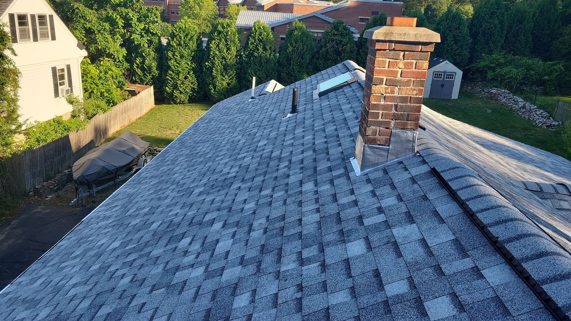 Gray shingle roof with a brick chimney and several vents on a sunny day.