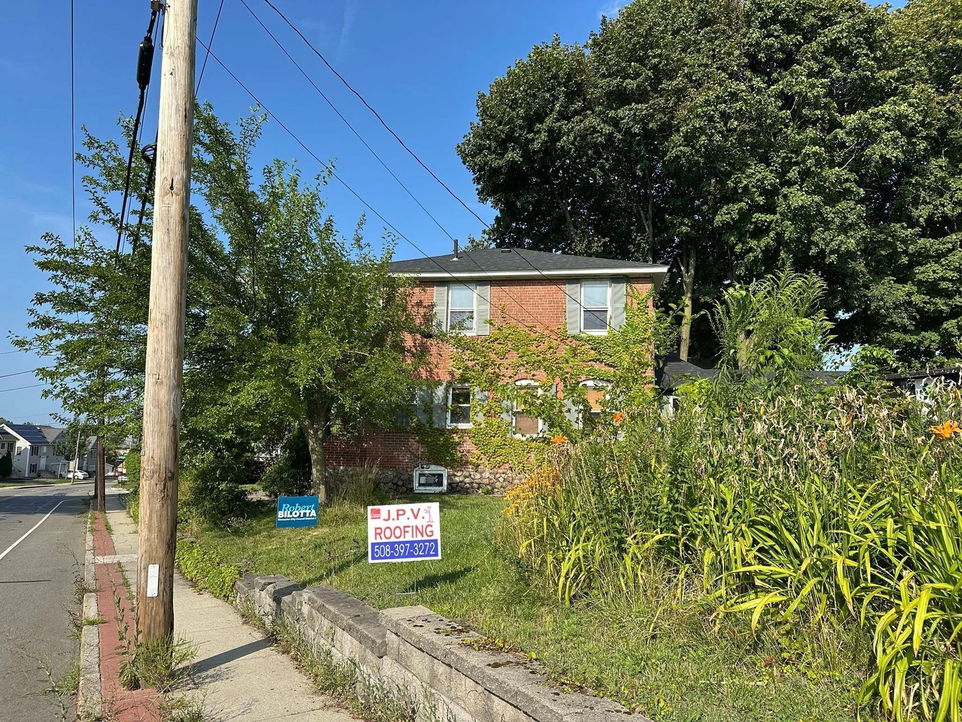 Two-story brick building with overgrown vegetation. Two signs in front. Sidewalk and road.