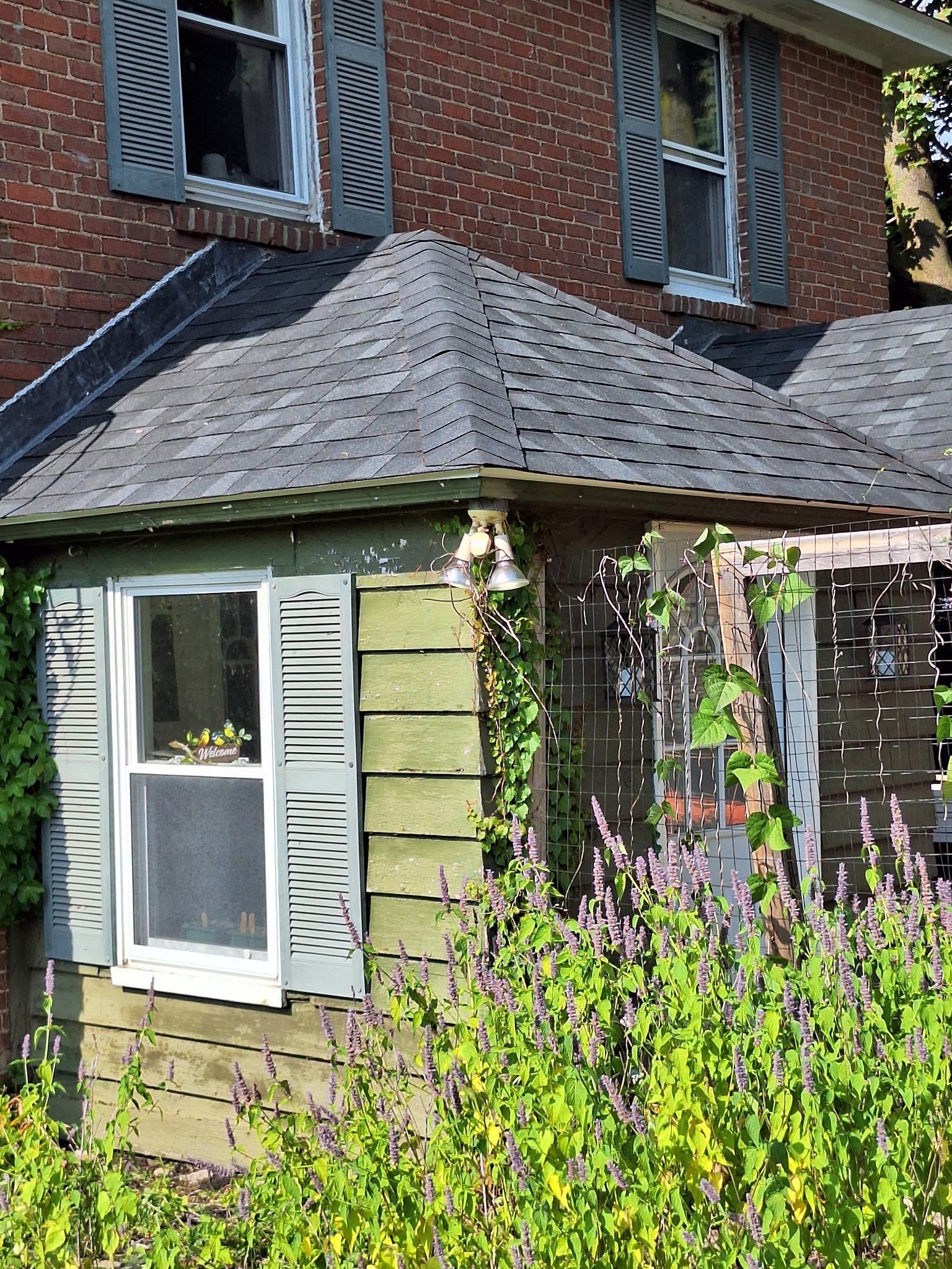 Brick house with dark shutters, covered porch, and overgrown plants.