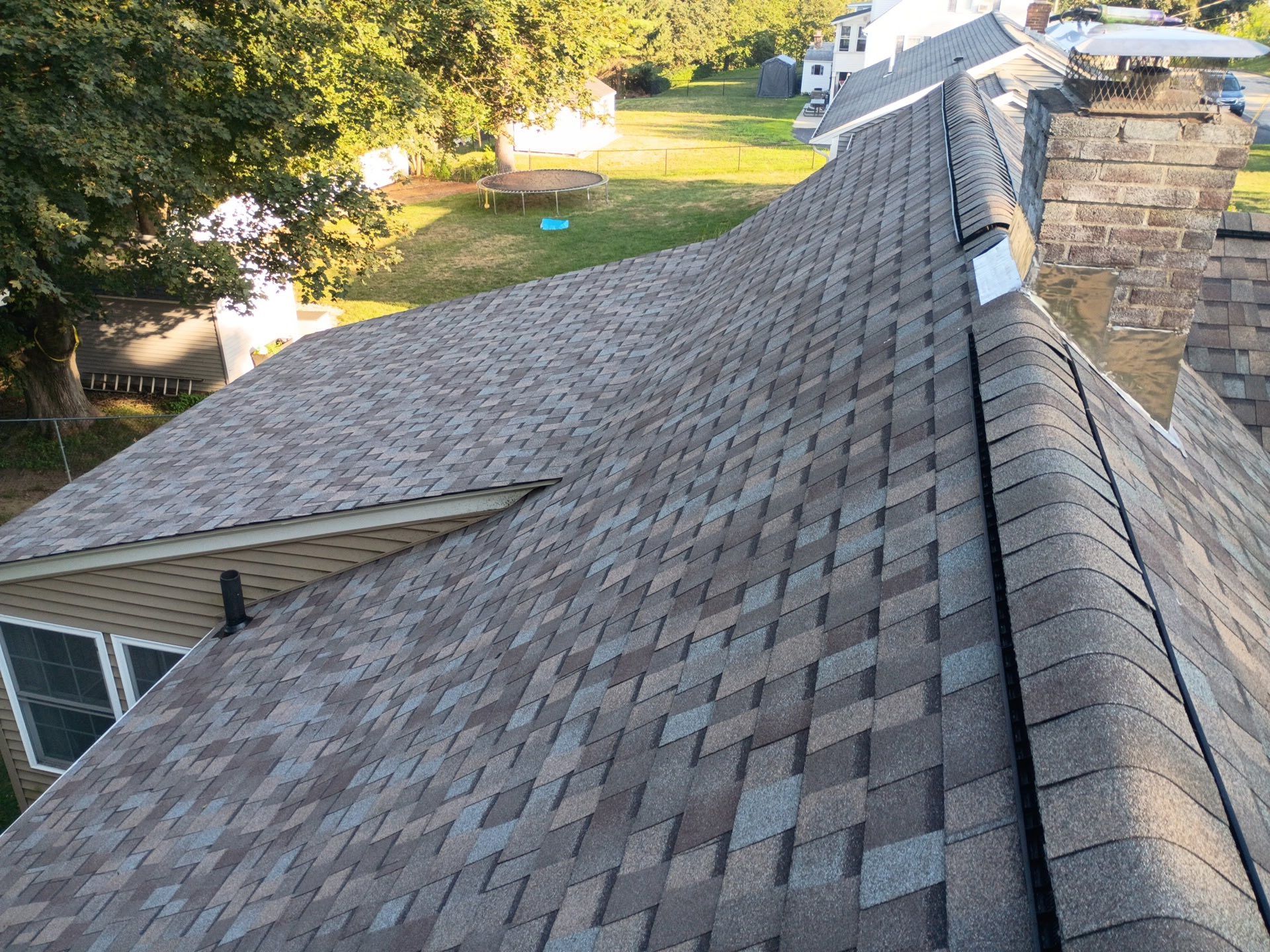 Shingled roof in shades of gray and brown on a house, chimney, and surrounding green trees and yard.