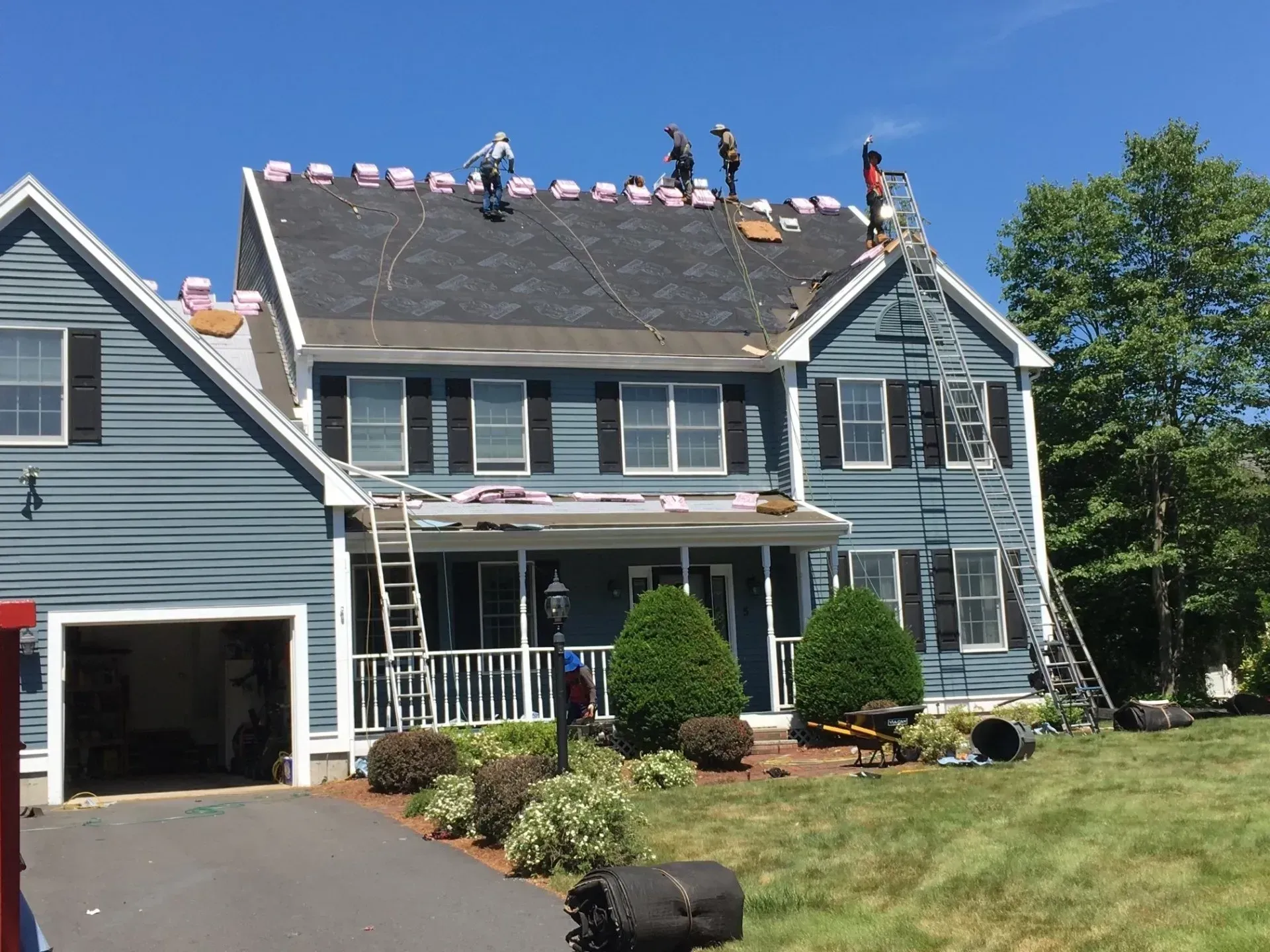 Roofers replacing roof on a two-story blue house; workers on roof and ladders, clear sky.