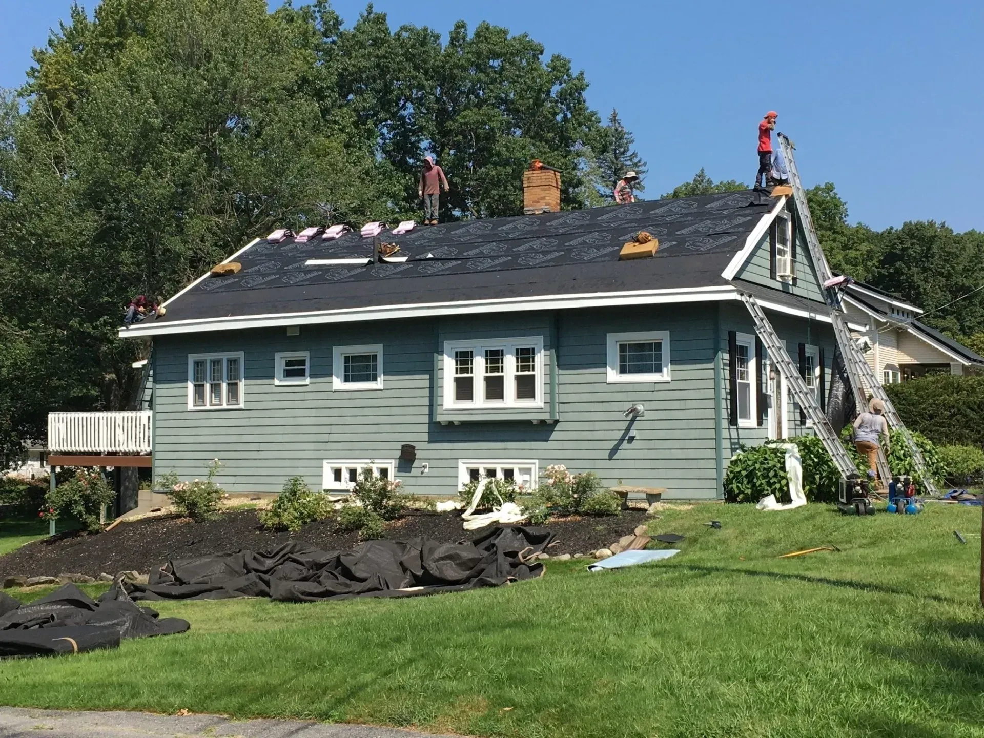 Workers replacing roof shingles on a light blue house with green grass and a sunny sky.
