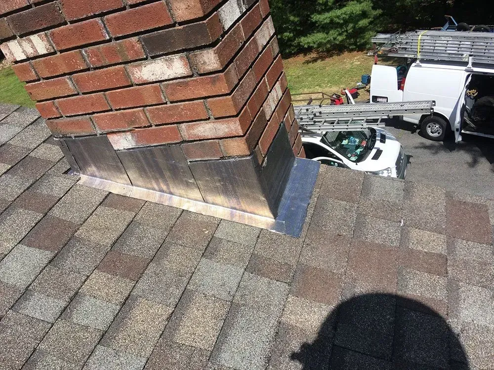 Brick chimney on a shingled roof with metal flashing; a white van and car are parked nearby.