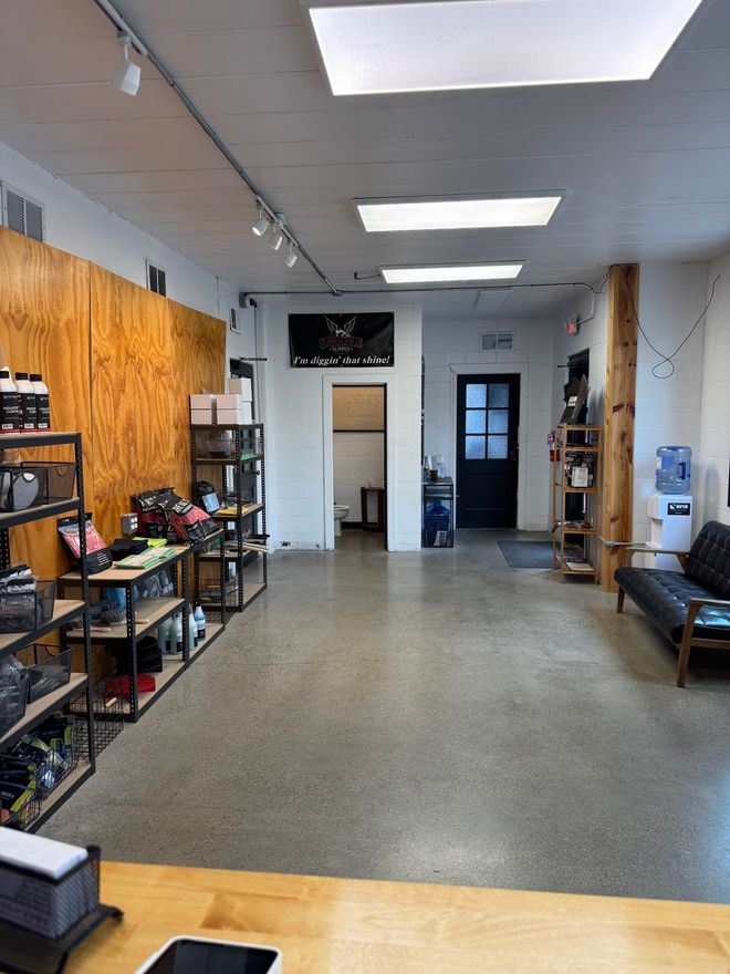 Long, narrow workshop or storage room with shelves, tables, tools, and a door at the back