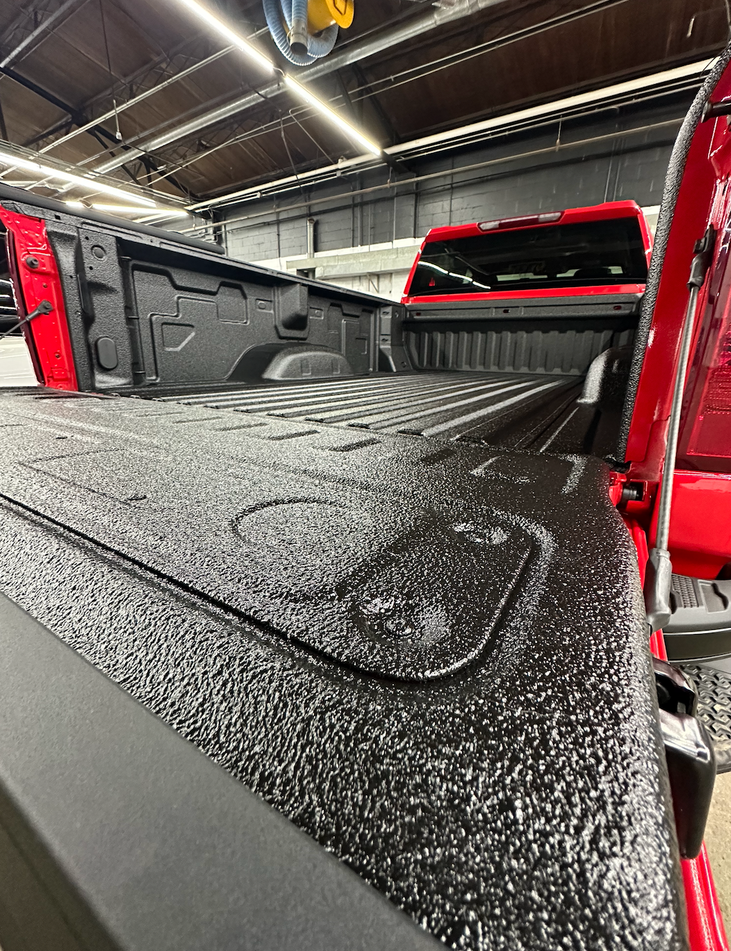Red pickup truck bed with black textured bed cover inside a garage or workshop.