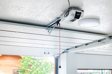 A ceiling-mounted automatic garage door opener with a red manual emergency release cord in a white garage.