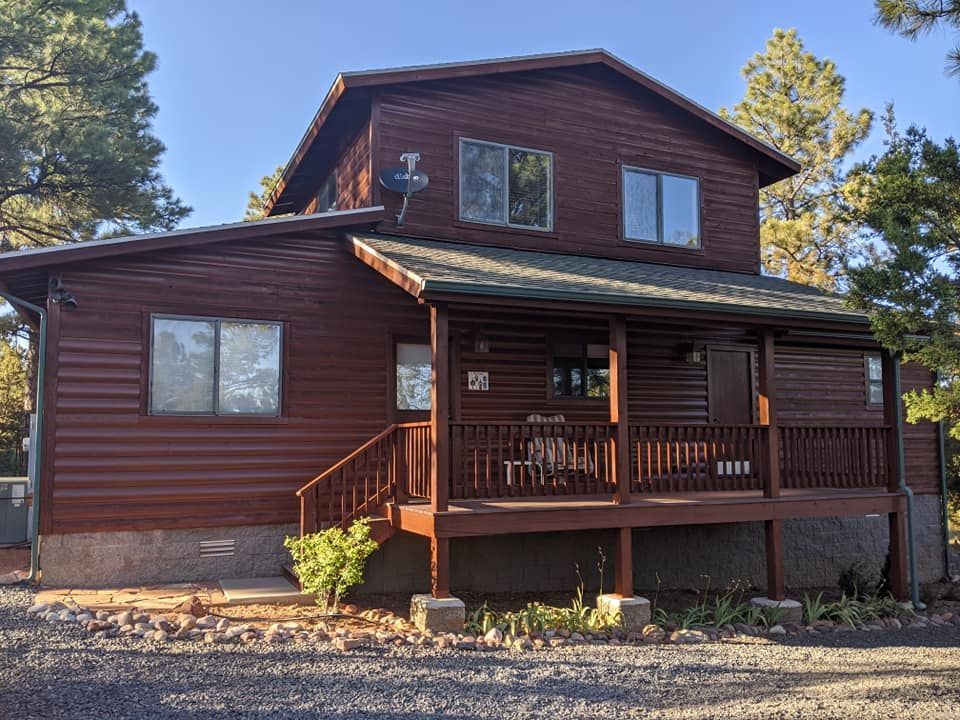 Two-story brown log cabin with porch, windows, and satellite dish in a wooded setting.