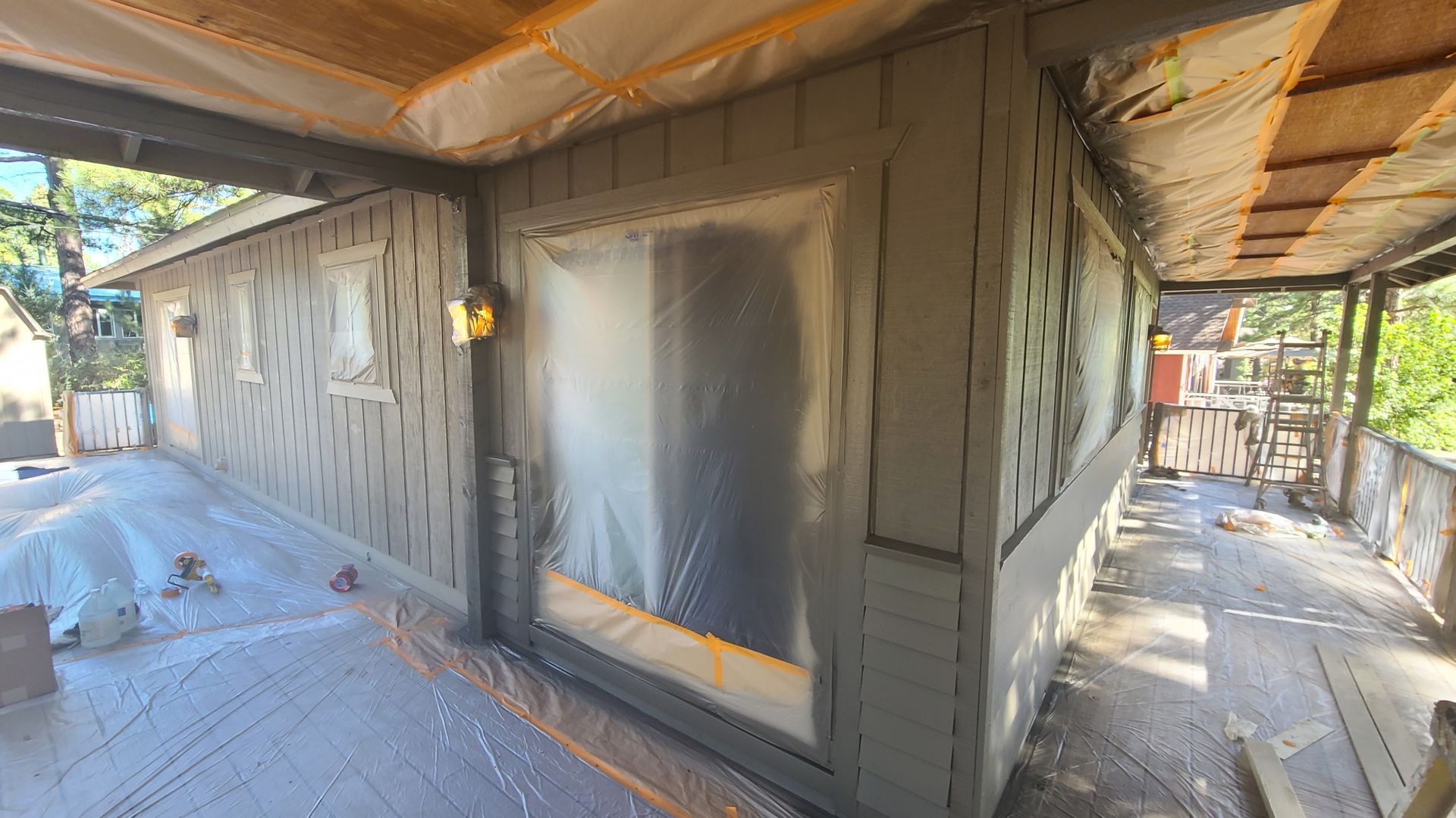 Exterior of a house being painted. Gray siding, plastic sheeting covering windows, under a porch.