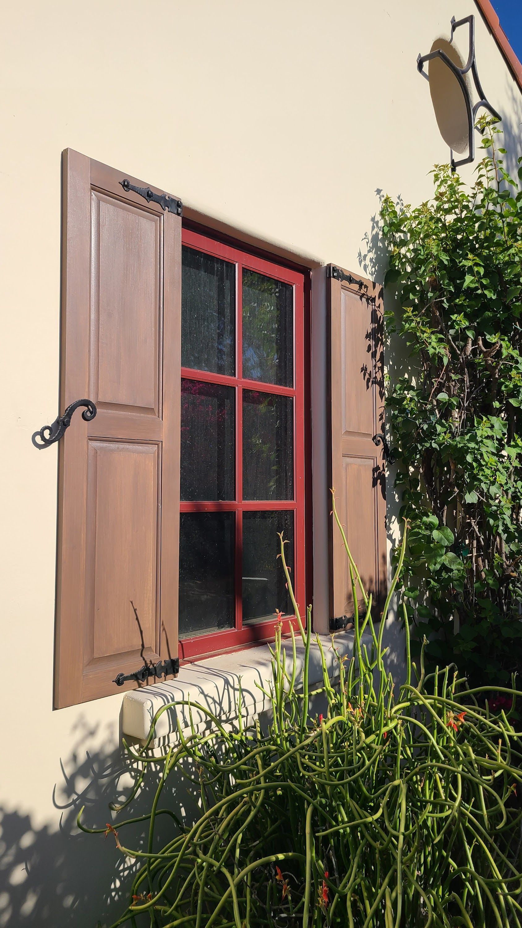 Window with brown shutters and red frame, on a beige wall next to green foliage.