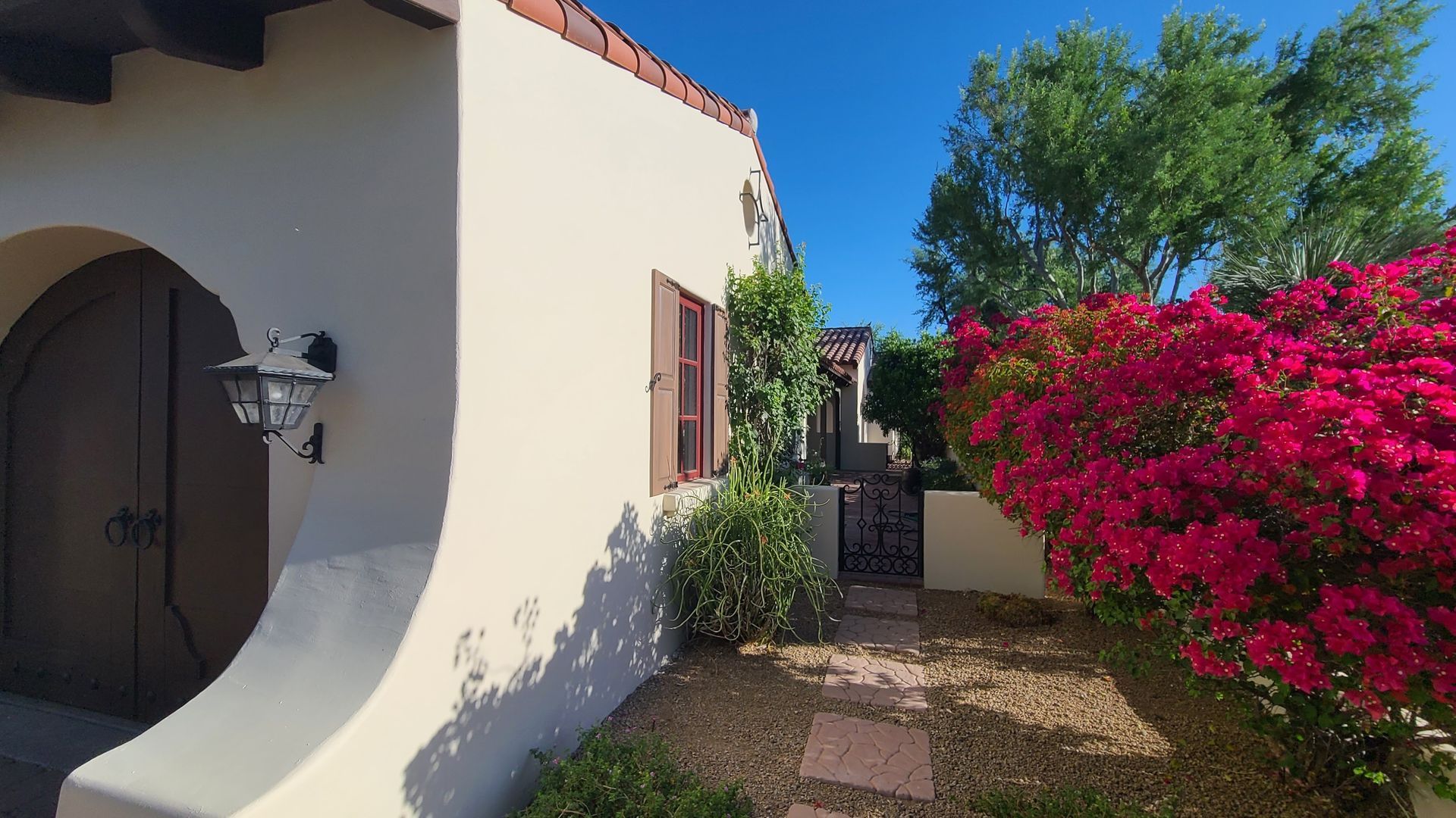 Tan stucco house with arched doorway, red bougainvillea bush, and blue sky.