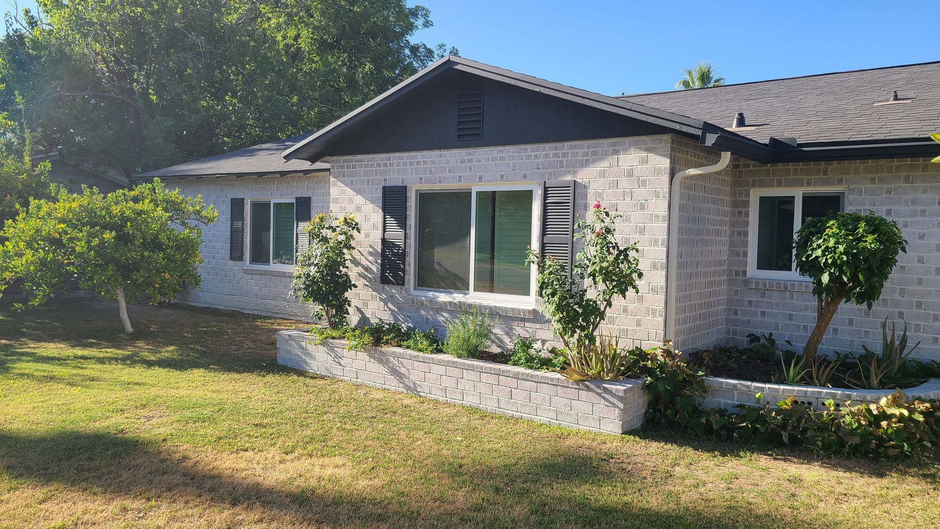 Brick house with black shutters and a small garden, set in a yard with a green lawn and trees.