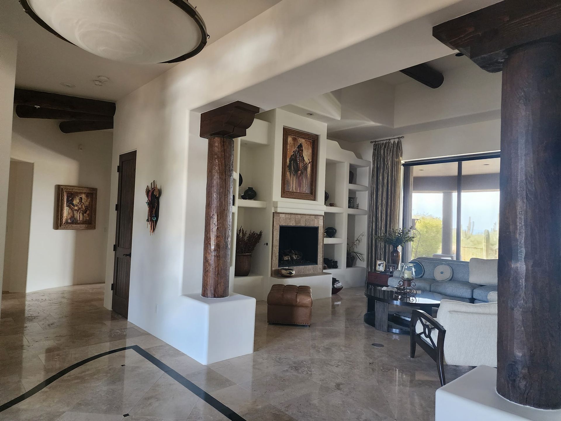 Spacious interior with stone floors, wooden pillars, fireplace, artwork, and a view through large windows.