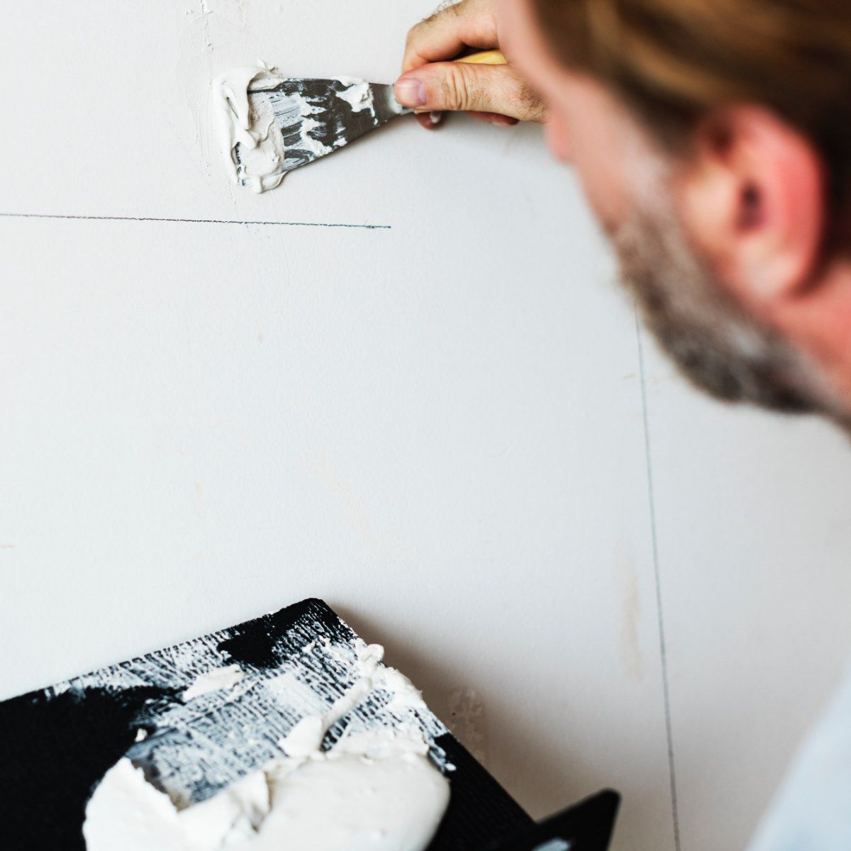 Person using a putty knife to apply spackle to a wall, with a tray of spackle in the foreground.