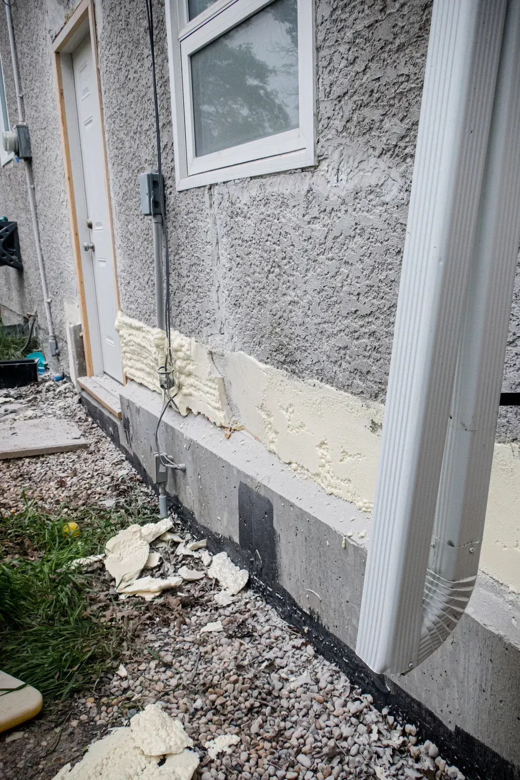 Exterior wall with foam insulation being applied to a concrete foundation. A door and window are visible.