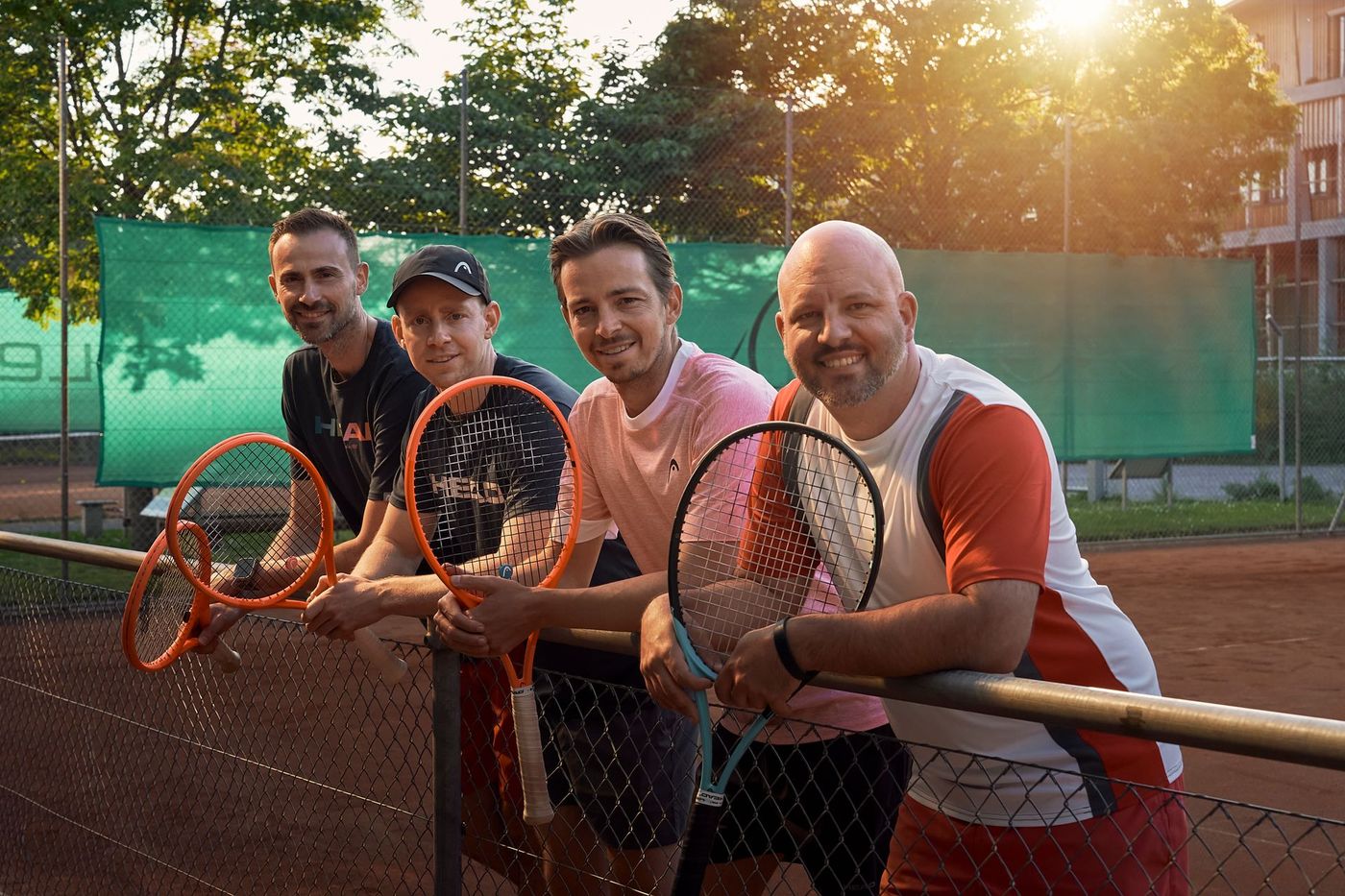 Eine Gruppe von Männern steht mit Tennisschlägern in den Händen nebeneinander auf einem Tennisplatz.