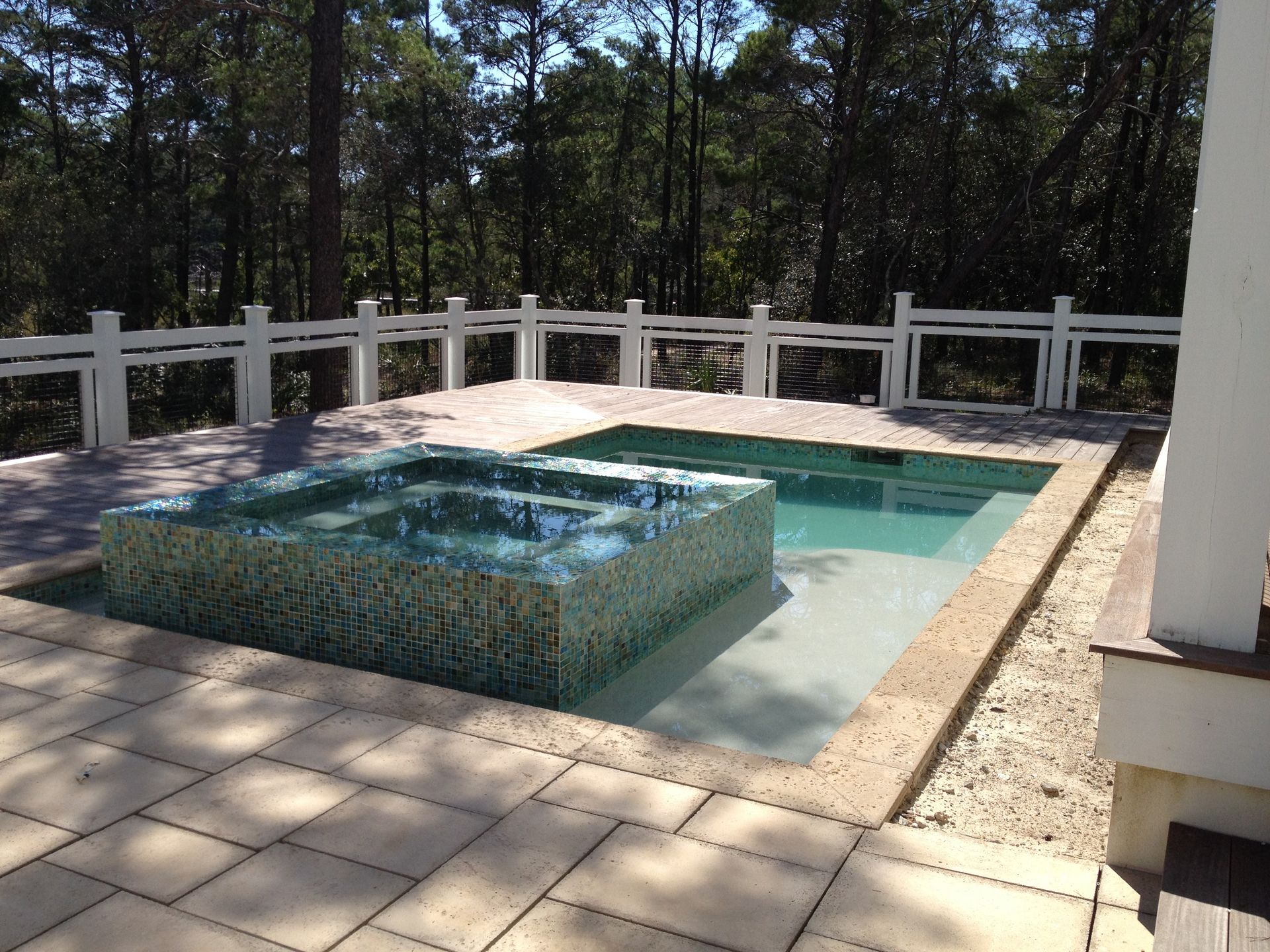 A square, tiled hot tub centered within a rectangular swimming pool on a patio deck, surrounded by a white picket fence.