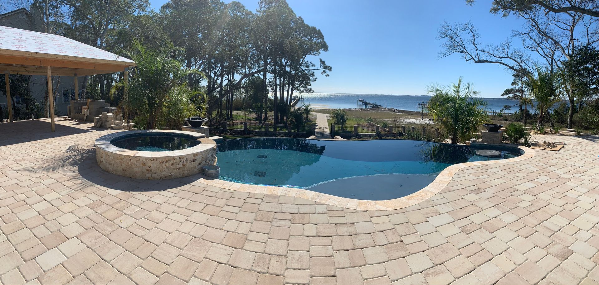 A backyard pool and hot tub on a stone patio overlooking a beach with trees under a clear blue sky.