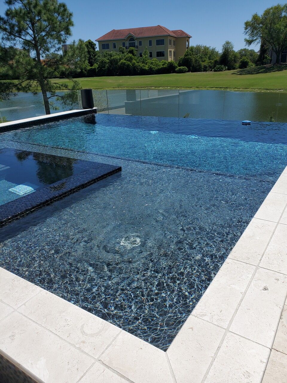 An infinity-edge swimming pool with dark blue textured tiles, overlooking a pond and house under a clear sky.