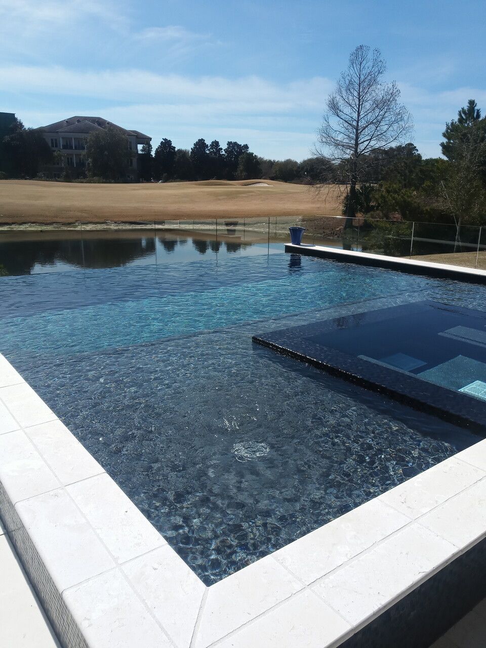 Pool with dark bottom, tiled edge, merging into a body of water with golf course and blue sky in background.