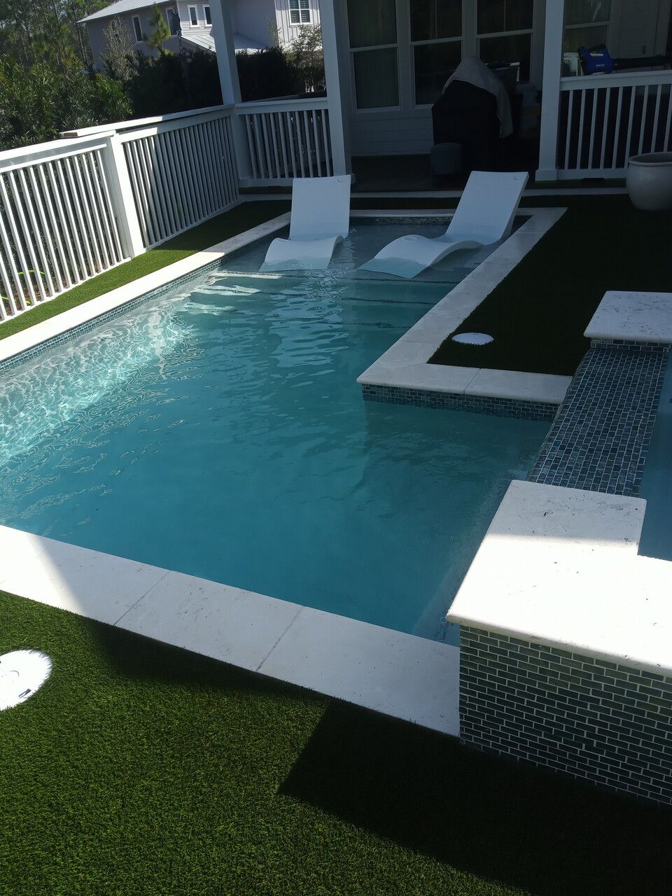A small, rectangular pool with blue water and two white submerged lounge chairs, set in a yard with artificial turf.