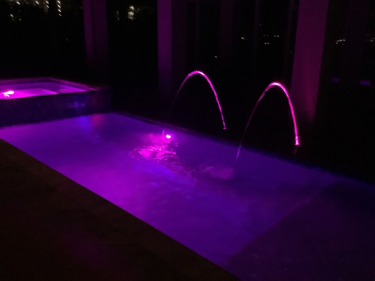 A swimming pool and hot tub at night, illuminated with vibrant purple underwater lighting and two arched water fountains.