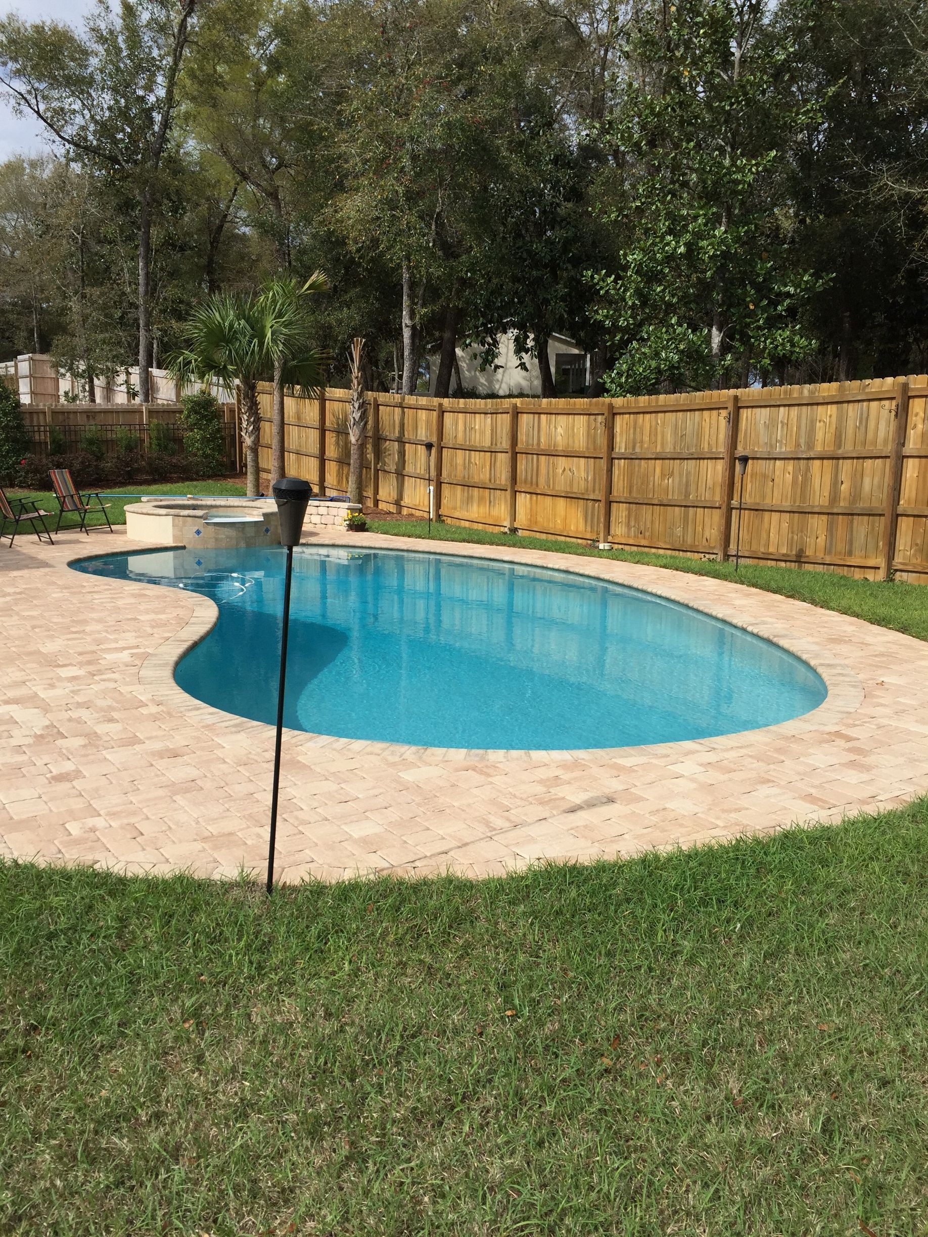A kidney-shaped swimming pool with a stone deck and a wooden privacy fence, surrounded by green grass and trees.