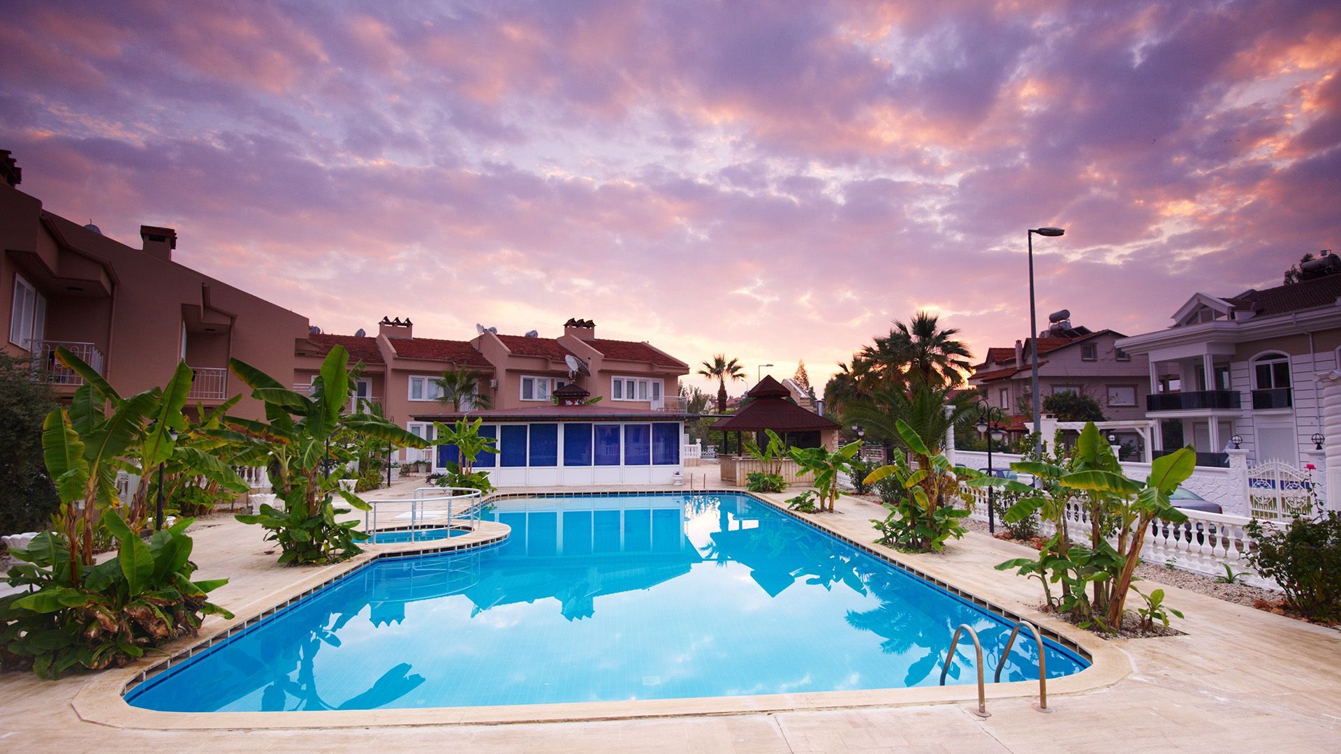 A swimming pool at a resort at sunset, surrounded by palm trees and apartment-style buildings under a purple sky.