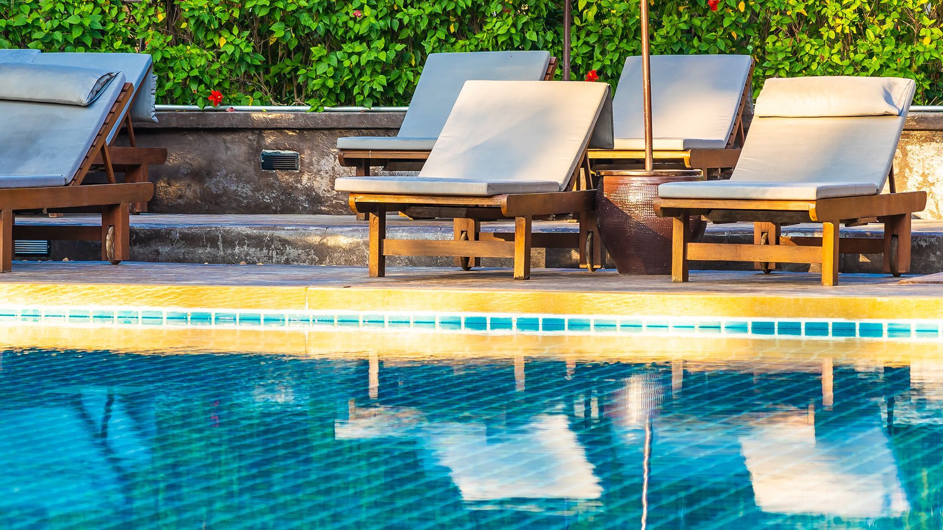 A sunny pool deck features several cushioned lounge chairs lined up beside a blue tiled swimming pool.