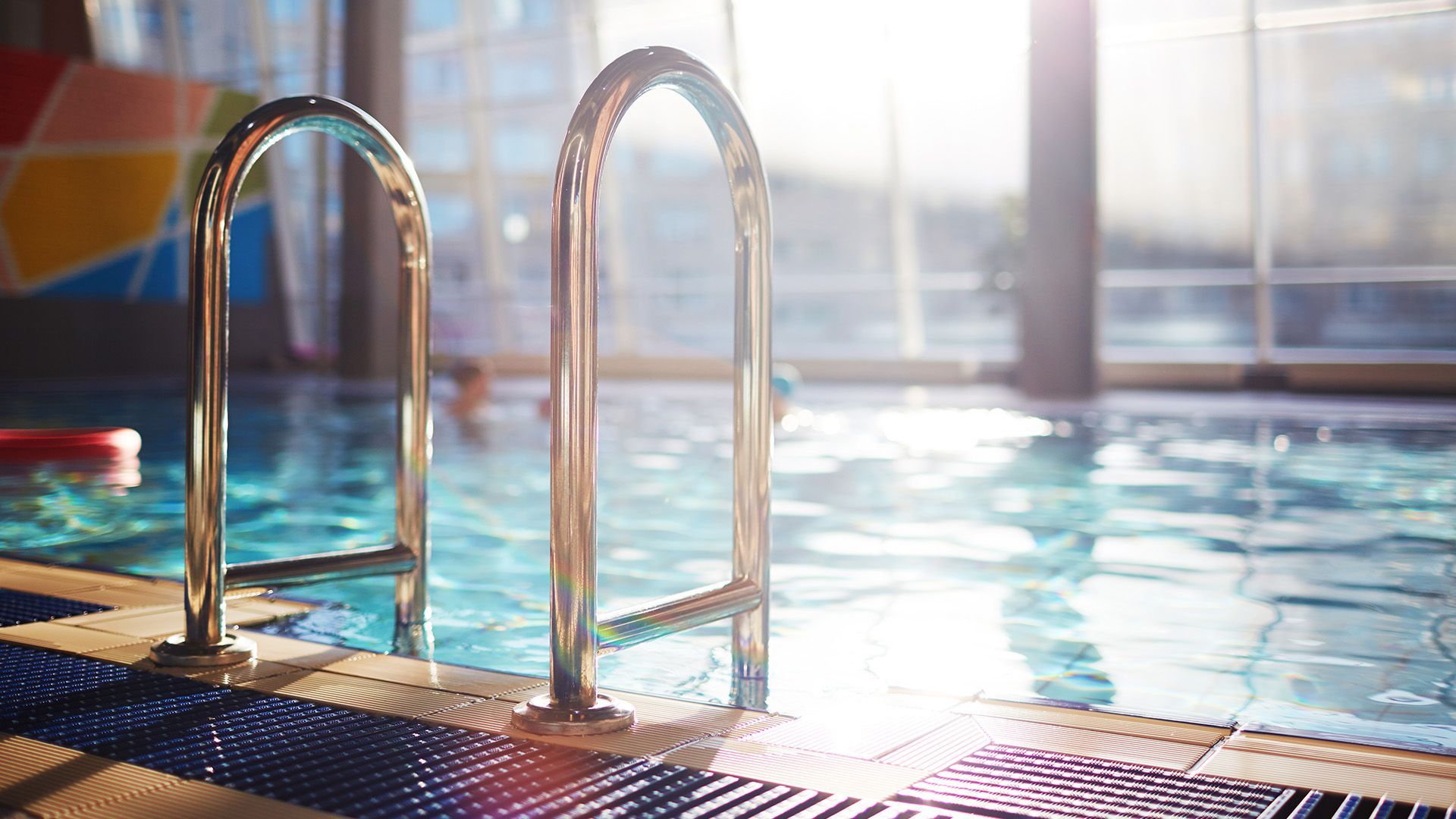 Metal pool ladders at the edge of an indoor swimming pool, with bright, sunlit water and blurred swimmers in the distance.