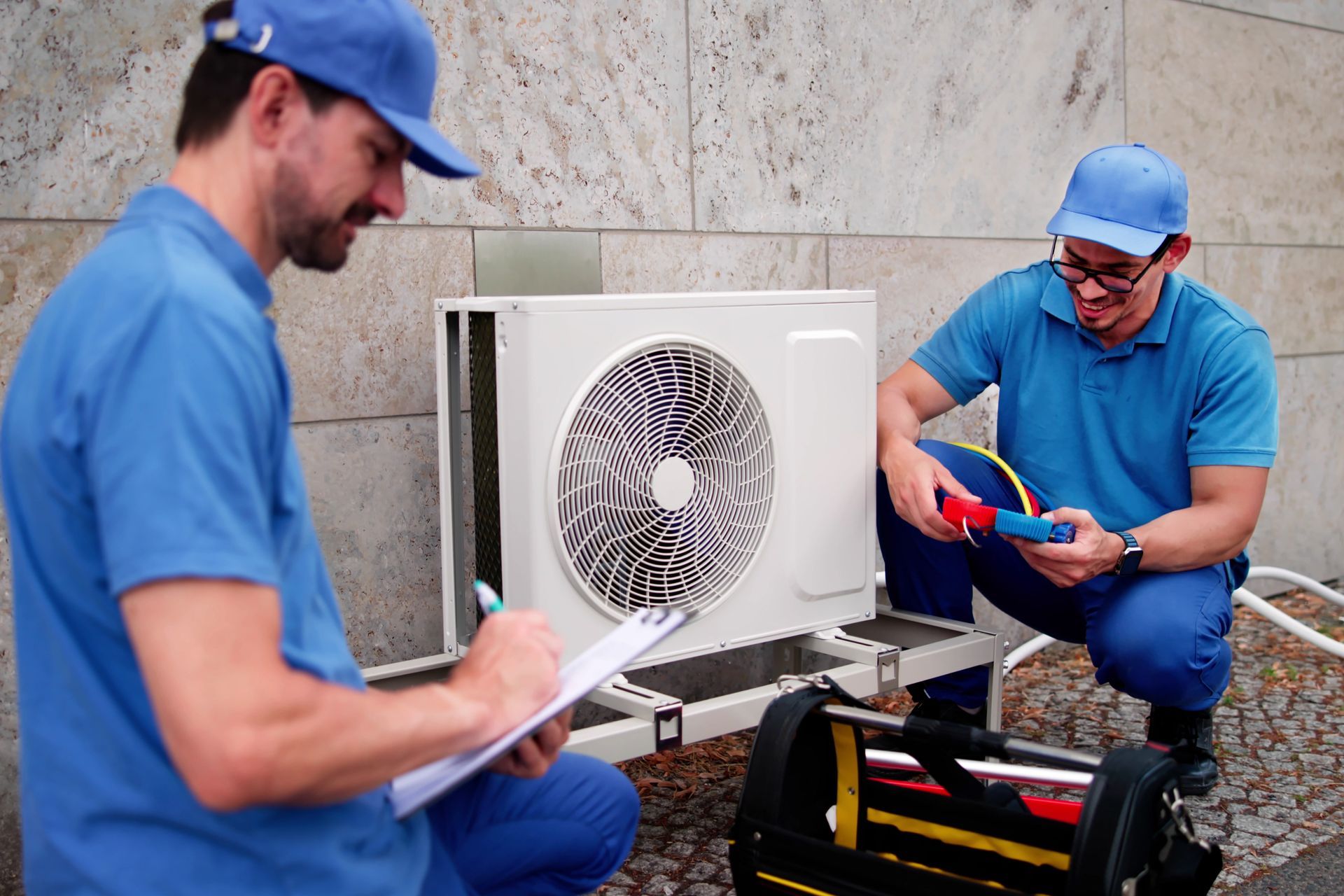 Two HVAC technicians inspecting an outdoor air conditioning unit. One writes on a clipboard, the other holds a tool.