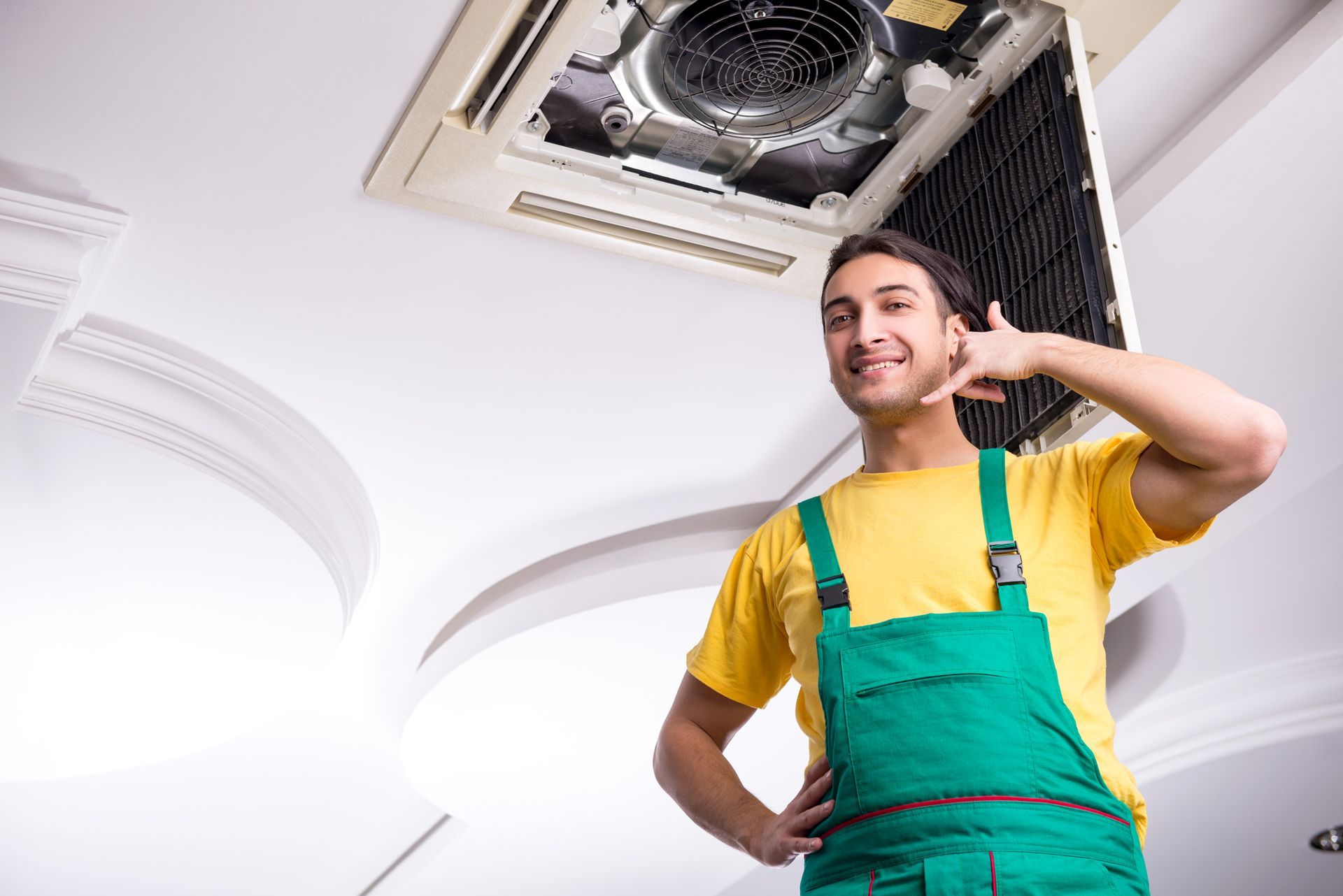 HVAC technician smiles, gesturing to call, standing below a ceiling air vent.