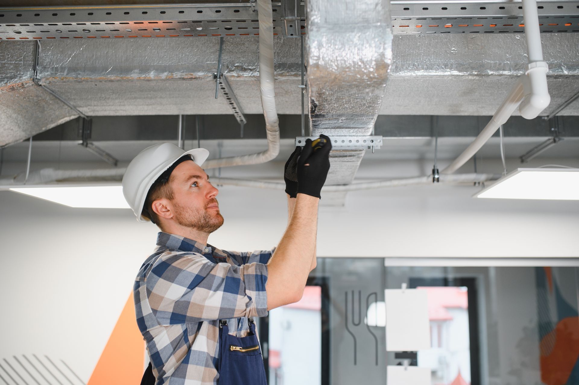 Construction worker in hard hat inspecting ductwork on a ceiling.