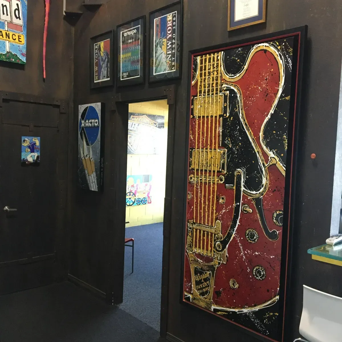 Entrance to a room with art. A gold and red guitar art piece hangs on the right. Doorway leads to another room with art.