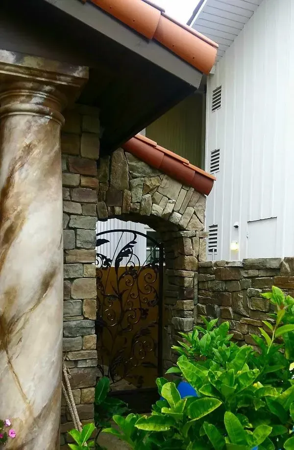 Stone archway with wrought iron gate, supported by marbled column and framed by green foliage.