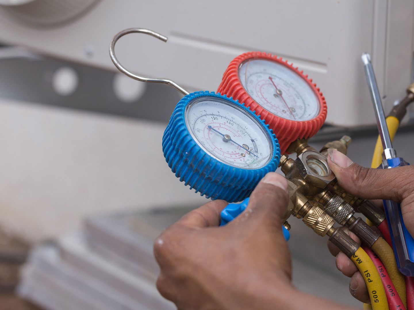 Hands adjusting gauges attached to an air conditioning unit. The gauges are blue and red, with hoses.