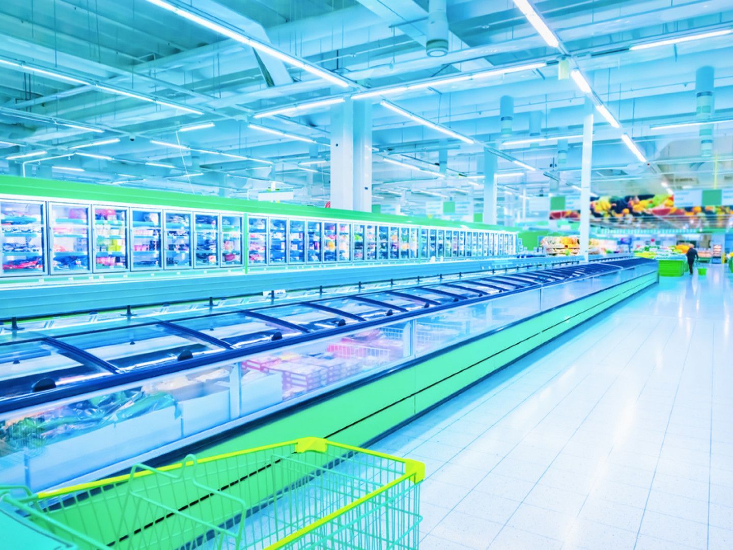 A brightly lit grocery store aisle with long freezers, cart, and a person in the distance.